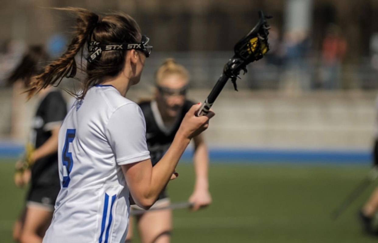 Female Identifying lacrosse student athlete on the field with lacrosse stick in hand, action shot